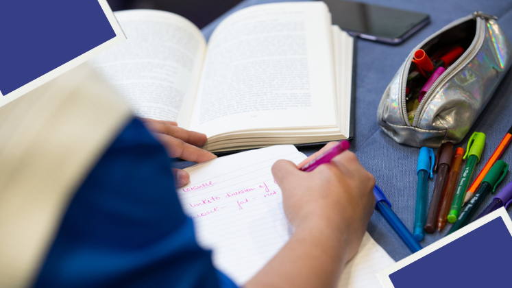 A person writes in a notebook with a book in front of them surrounded by coloured pens