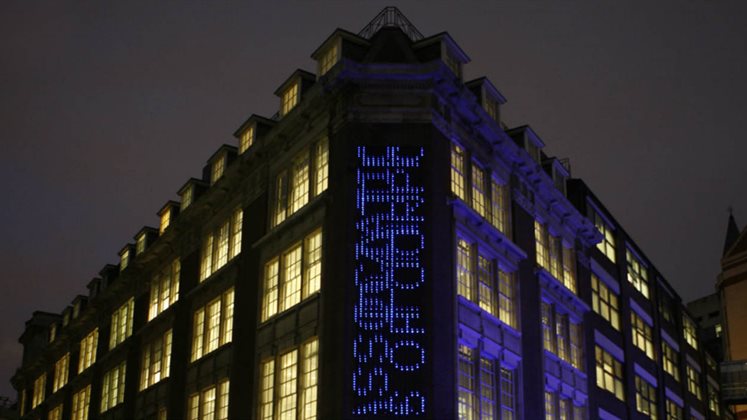 An outside view of LSE Library at night which includes the artwork Bluerain - an installation where searches from the catalogue scroll down from top to bottom on top of each other using blue LED lights.