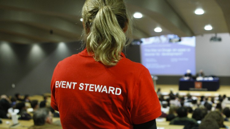 A person is stood wearing an red t-shirt with 'Event Steward' written on the back. They are looking out over the audience of an event.