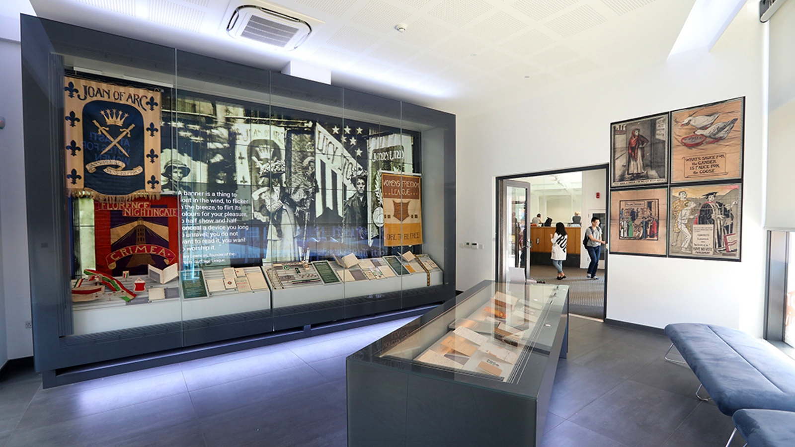 A view of the Exhibition Gallery looking primarily at the large digital screen where images from the exhibition along with various objects are on display. It also includes the main display case with objects in and a view out of the room's door into LSE Library.