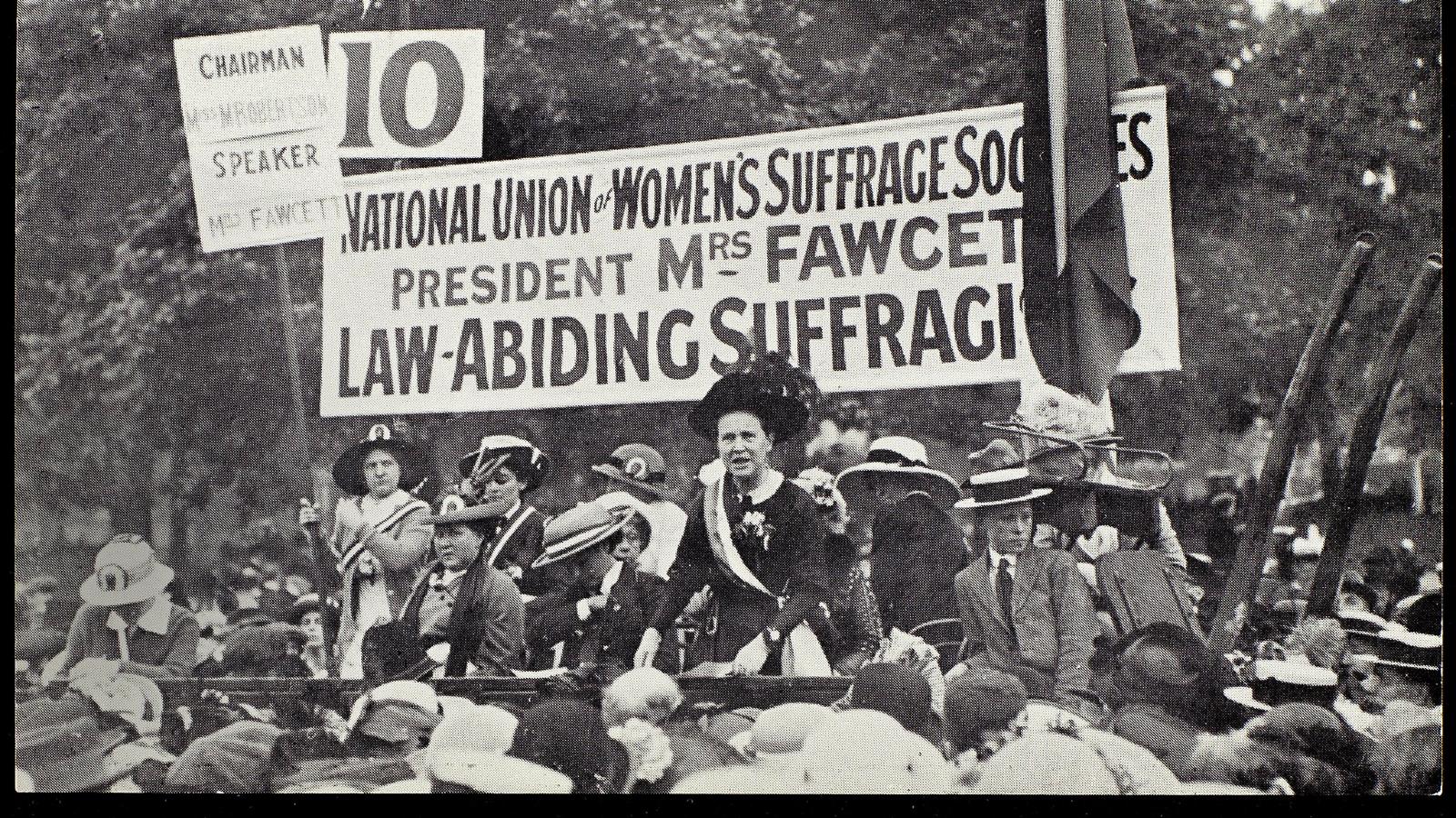 A group of people with banners and placards at a protest looking into the camera. One of them is Millicent Garrett Fawcett who is speaking at this protest as indicated by the banner above her head.