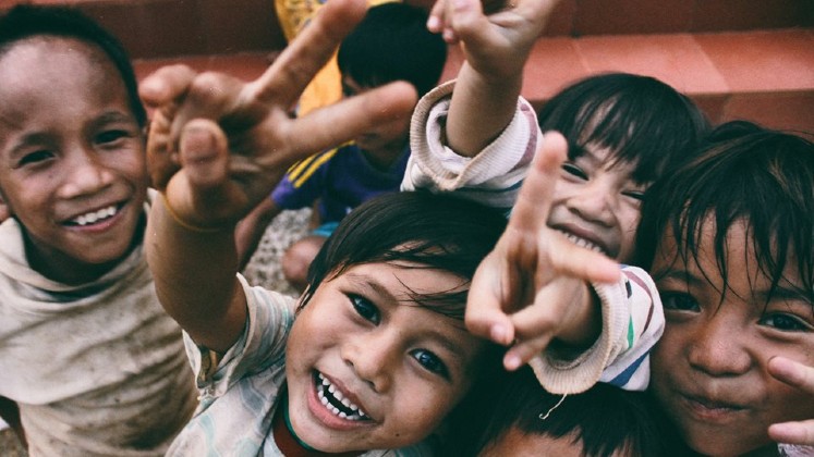 A group of smiling children all looking at the camera. Some are giving peace signs with their fingers