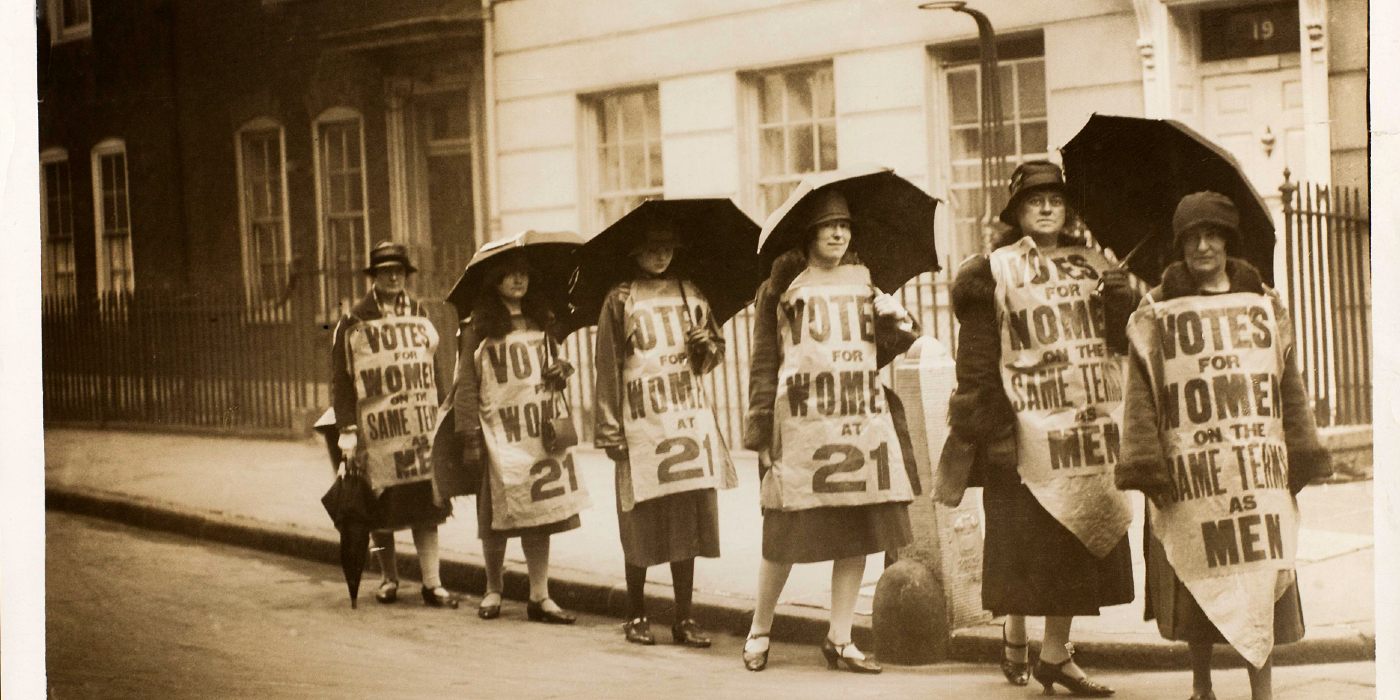 A group of women marching down a street with banners attached to them and carrying umbrellas.