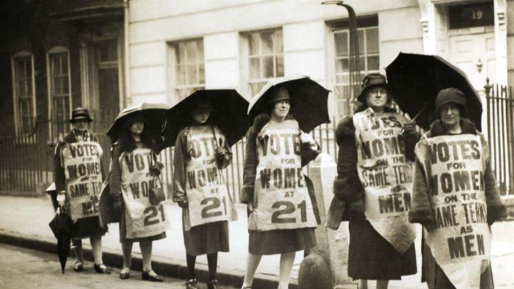 A group of women marching down a street with banners attached to them and carrying umbrellas.