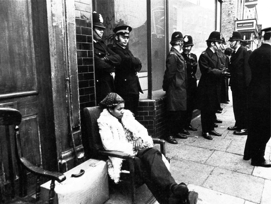 A person sat outside a building with belongings. Many police officers stood around the building.