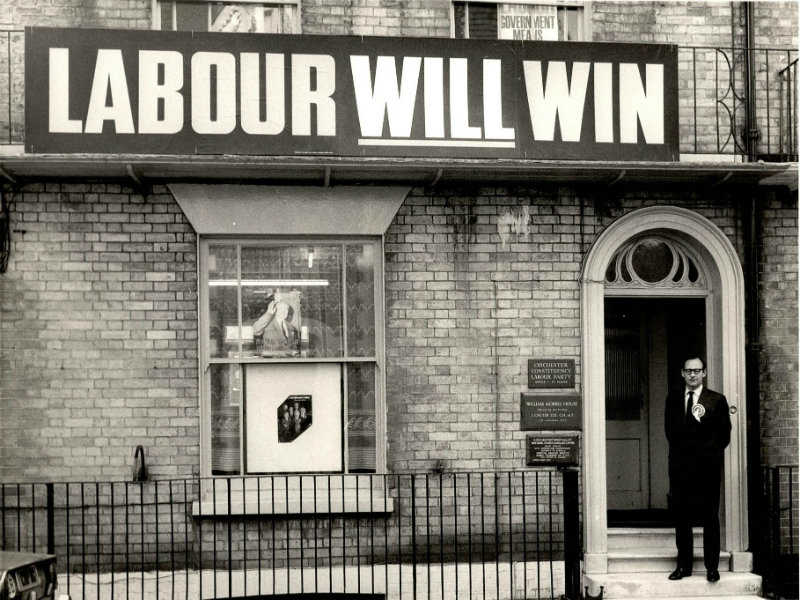 Man stood outside a house with a large 'Labour Will Win' banner on house front.