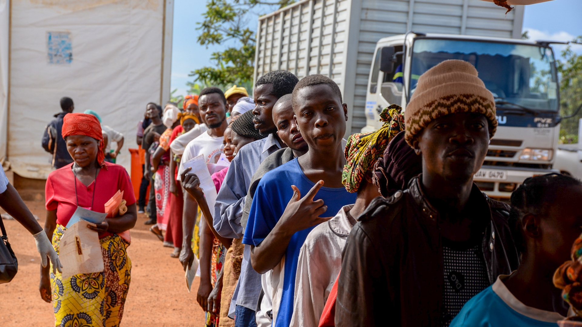 Group of African men, women and children in a line with a truck in the background.